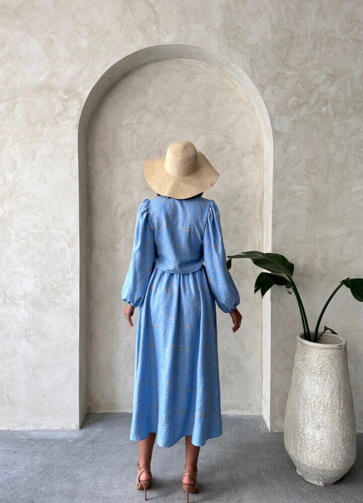 Model wearing a blue boho printed linen two-piece set standing in an archway with a potted plant.