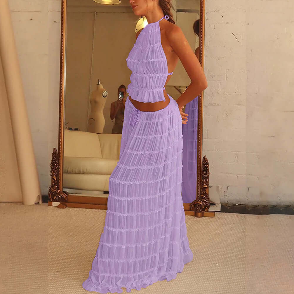 A side view of a woman wearing a light purple, two-piece outfit. The outfit consists of a ruffled crop top with a halter neckline and a matching long, pleated skirt. The woman is standing indoors, with a large mirror  in the background.