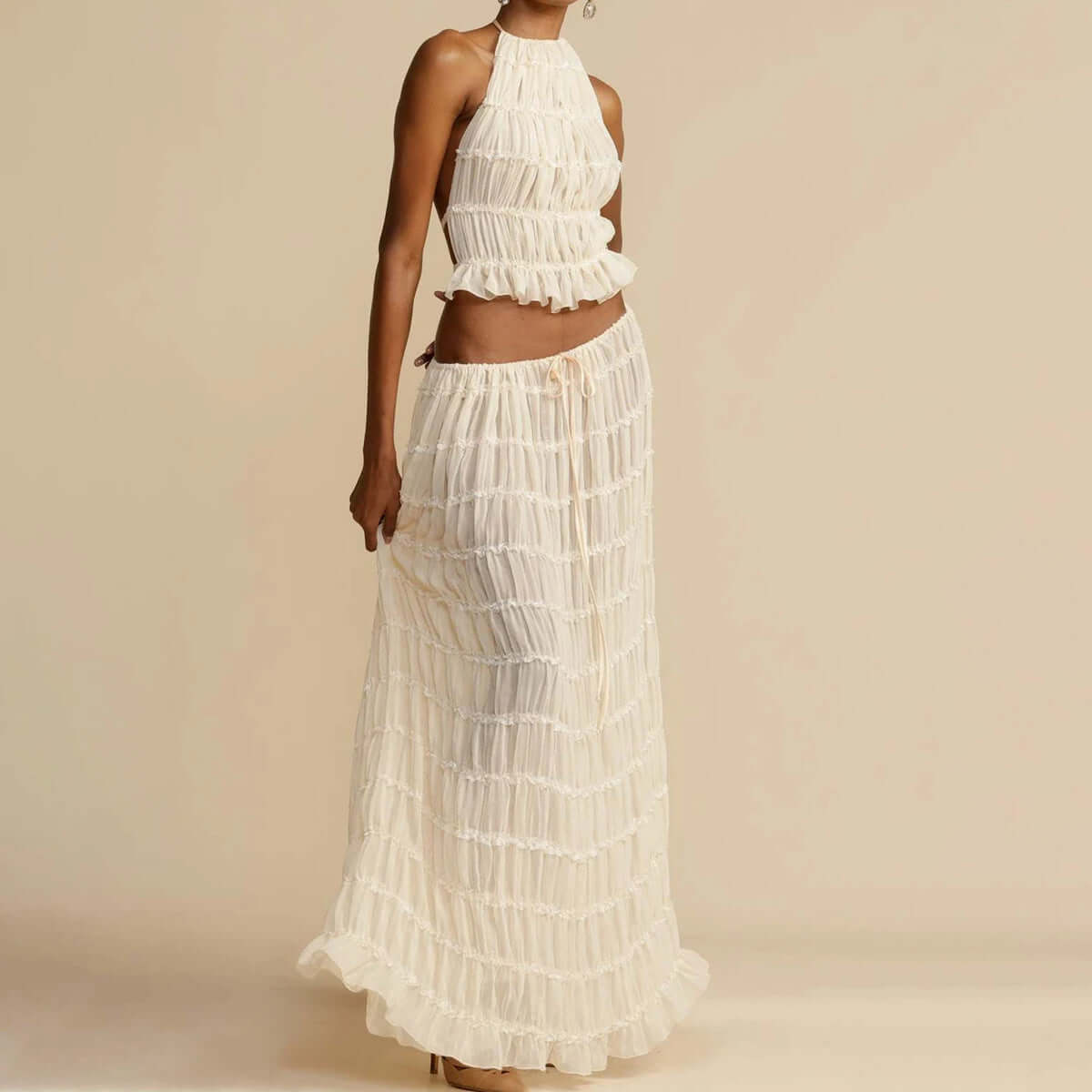 A full-length view of a woman wearing an off-white two-piece outfit. The fashionable ensemble features a ruffled halter-neck crop top and a matching long, tiered maxi skirt. The woman is standing on a light-colored surface against a neutral background, showcasing this elegant women's wear.