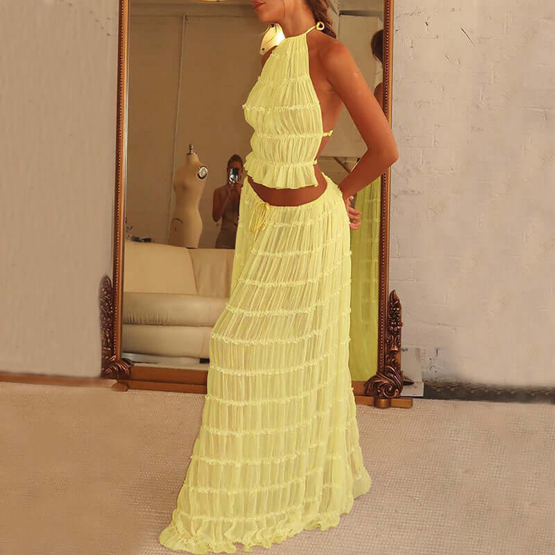 A side view of a woman wearing a light YELLOW, two-piece outfit. The outfit consists of a ruffled crop top with a halter neckline and a matching long, pleated skirt. The woman is standing indoors, with a large mirror and what appears to be a dark wooden wall or furniture in the background.