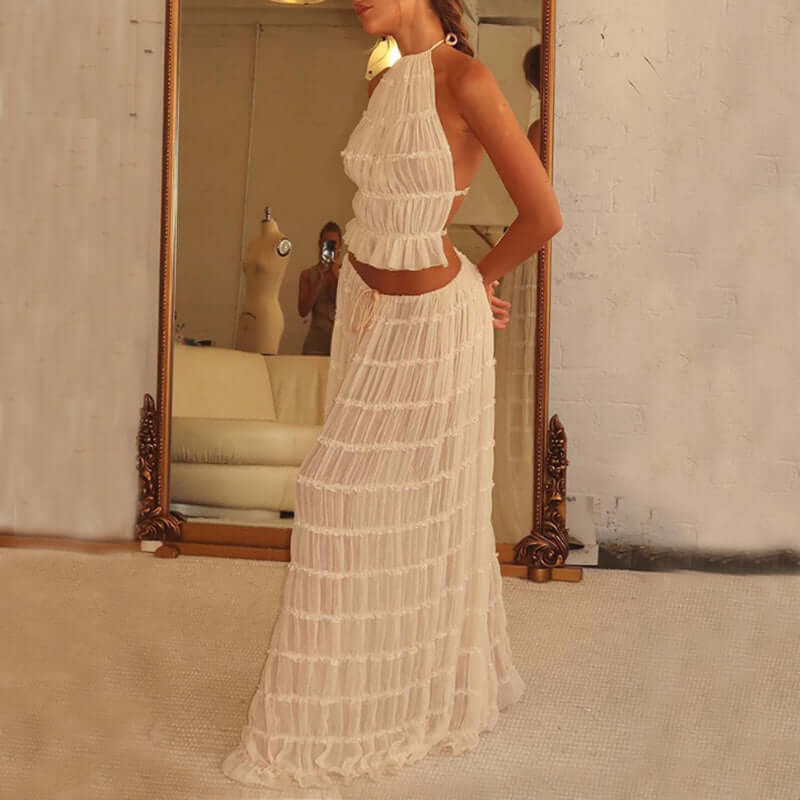 
A side view of a woman wearing a white, two-piece outfit consisting of a ruffled crop top and a matching long, tiered skirt. The woman is standing indoors, with a large decorative mirror and part of a plant visible in the background