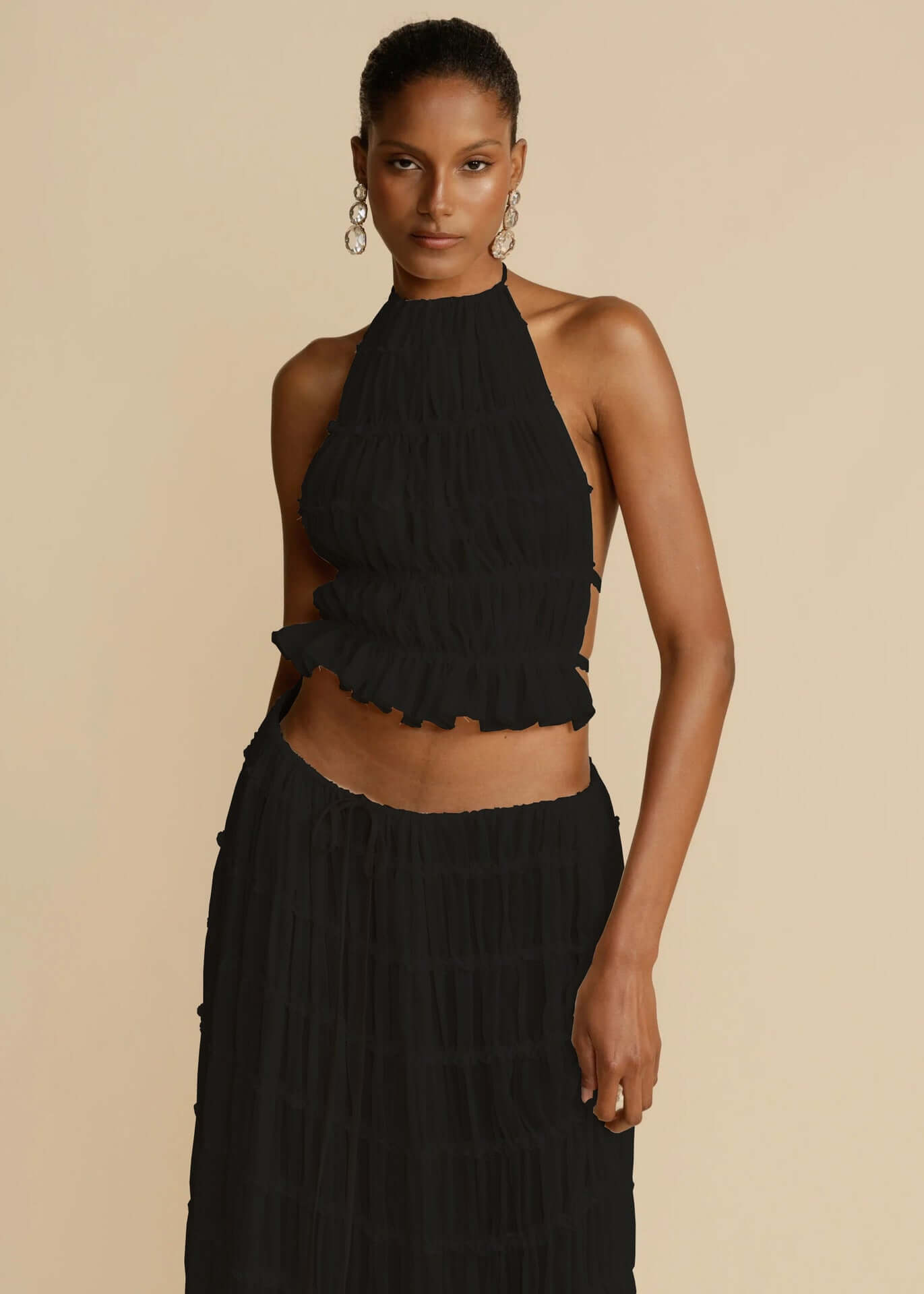 A front view of a woman wearing a black, two-piece outfit. The outfit includes a fitted, halter-neck crop top with a ruffled hem and a long, pleated black skirt. The woman has dark hair pulled back and is looking directly at the camera with a neutral expression. The background is solid 