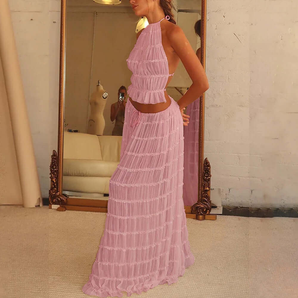 A side view of a woman wearing a pink, two-piece outfit. The outfit consists of a ruffled crop top with a halter neckline and a matching long, pleated skirt. The woman is standing indoors, with a large mirror in the background.