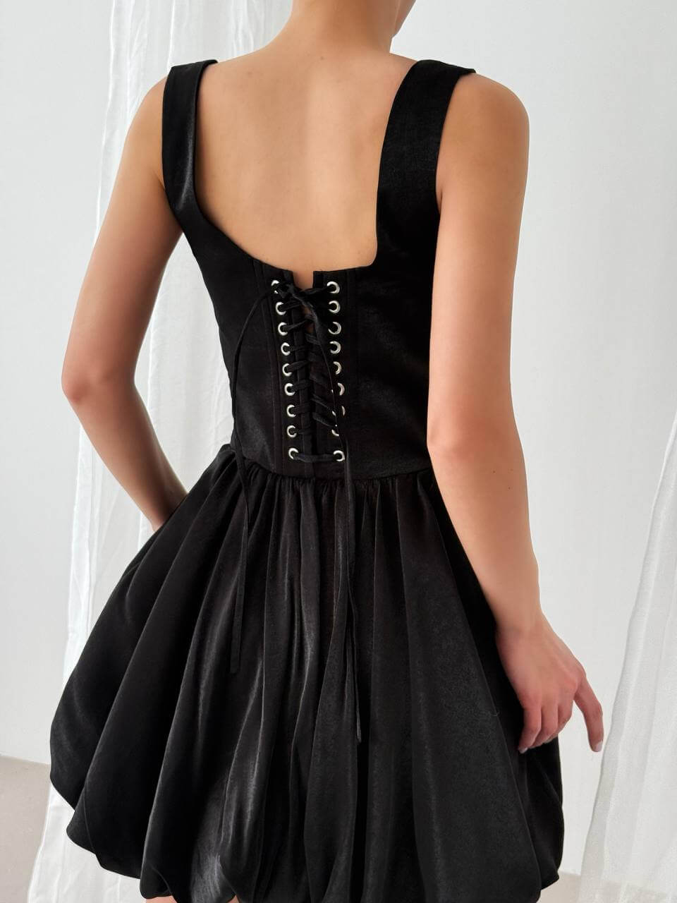 The back of a sleeveless black mini dress featuring a corset-style lace-up closure on the upper back and a gathered skirt.