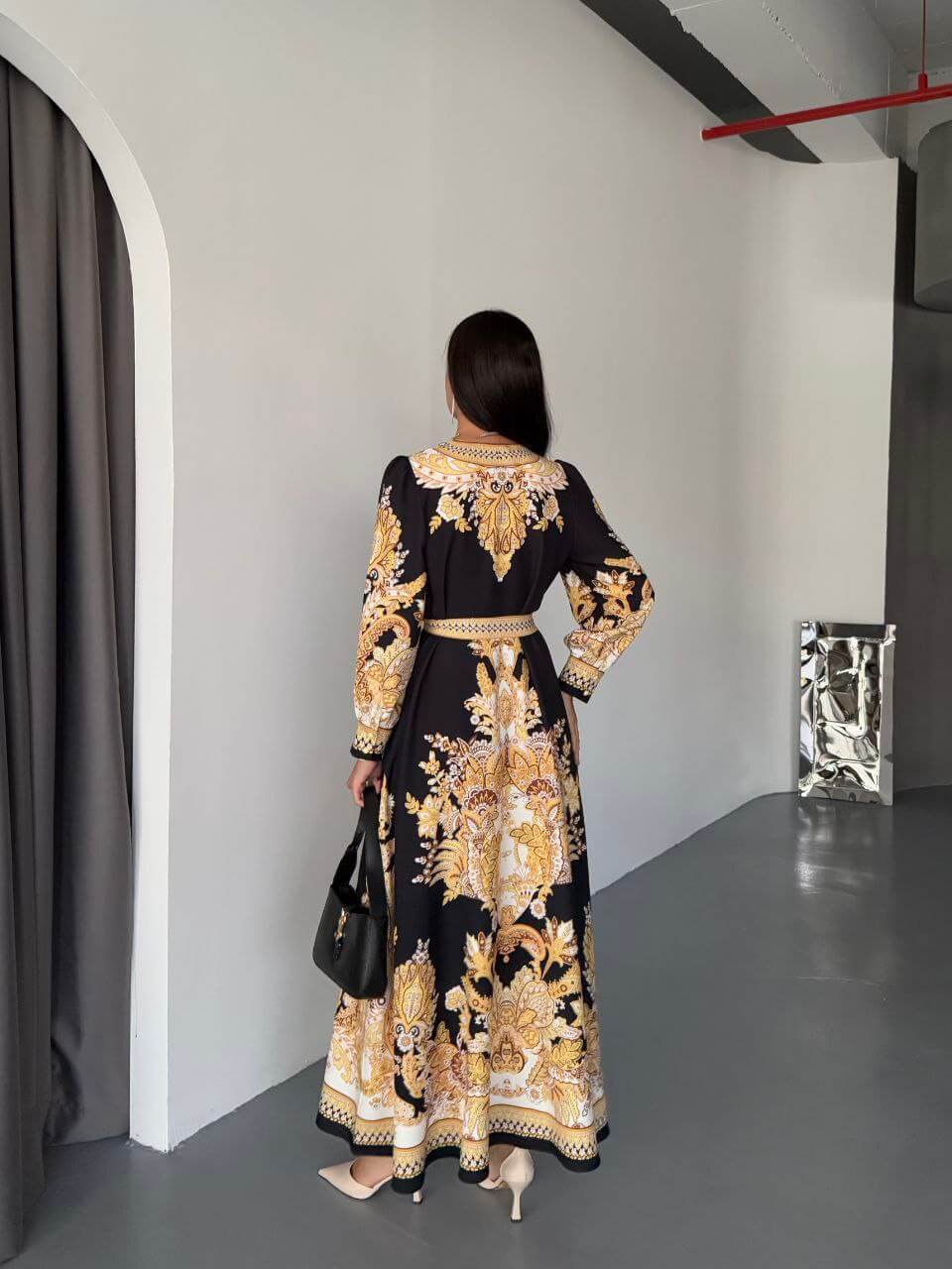 A woman standing with her back to the camera, wearing a long black dress with a stylish pattern on the fabric, and holding a bag.