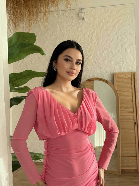 Model in a pink ruched long-sleeved dress with deep V-neckline, styled elegantly against a plant backdrop.