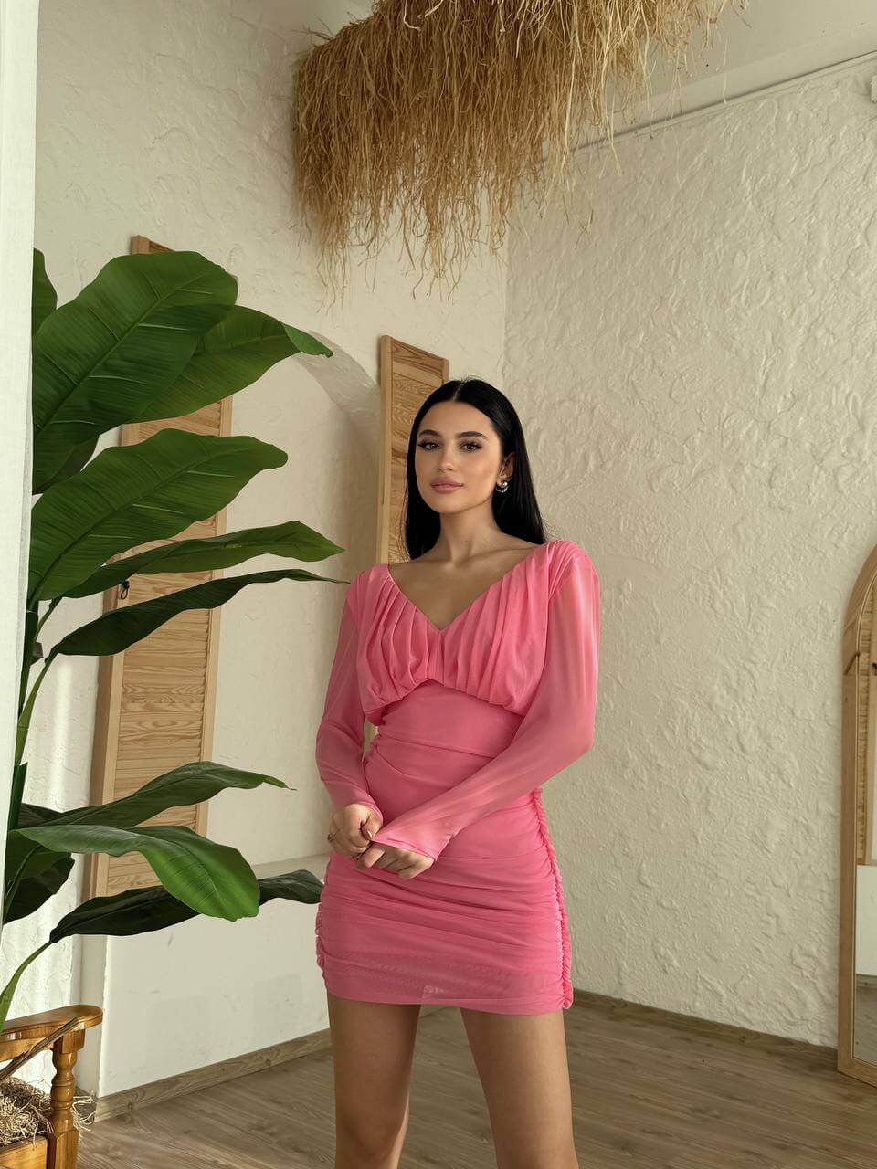Model wearing a pink ruched long-sleeved dress with a deep V-neckline against a textured wall.